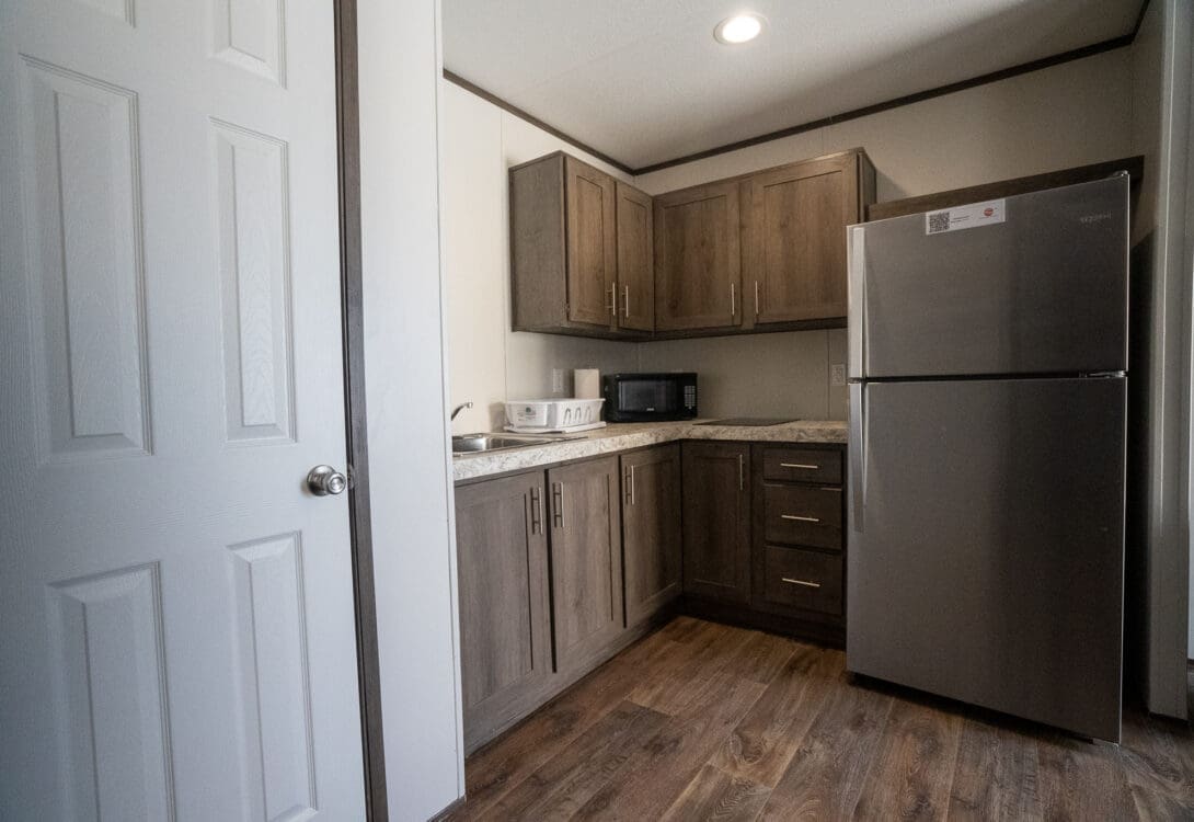A view of the kitchen area in CHH Pecos with gray two-door refrigerator, microwave and kitchenwares on countertop, brown cabinets and wooden floor